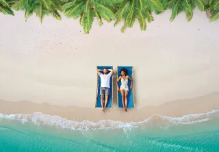 Couple se relaxant sur des transats au bord d’une plage de sable fin bordée de palmiers, face à la mer turquoise des Caraïbes