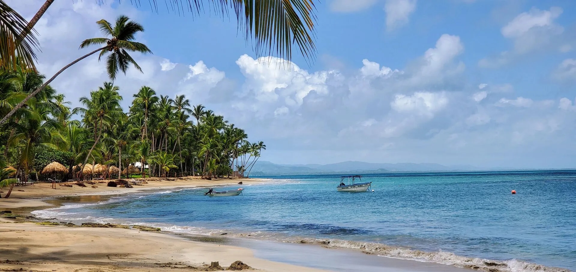 Plage de sable fin bordée de cocotiers avec bateaux au mouillage.