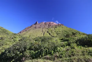 Vue en contre-plongée du volcan de la Soufrière en Guadeloupe sous un ciel bleu pur, montrant ses flancs escarpés recouverts d'une végétation basse et sa silhouette culminant à 1467 mètres d'altitude.