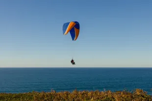 Baptême de parapente en tandem au-dessus de l'océan Atlantique en Guadeloupe, avec une aile bleue et orange sur fond de ciel clair et horizon marin.