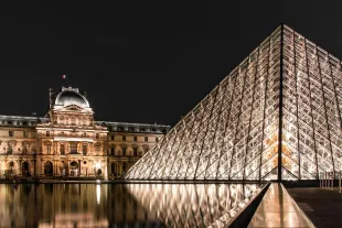 Vue nocturne de la Pyramide du Louvre illuminée avec le Palais du Louvre en arrière-plan, reflétés sur les bassins d'eau sous un ciel de nuit.