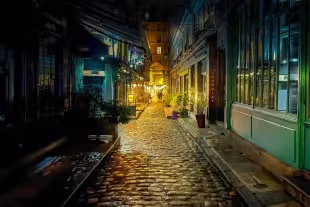 Rue pavée typique du quartier latin à Paris de nuit, illuminée par les lumières dorées des vitrines de commerces et de restaurants, avec la façade d'une chocolaterie au premier plan.