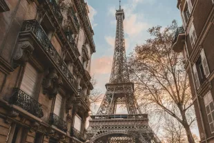 Vue en contre-plongée de la Tour Eiffel à Paris, s élevant majestueusement entre les façades haussmanniennes typiques de la capitale sous un ciel de fin de journée aux teintes orangées.