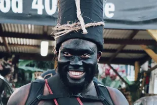 Portrait d'un membre d'un "groupe à po" au Carnaval de Guadeloupe, le visage enduit de noir de fumée et arborant une coiffe traditionnelle en tissu noir ornée de fibres naturelles.