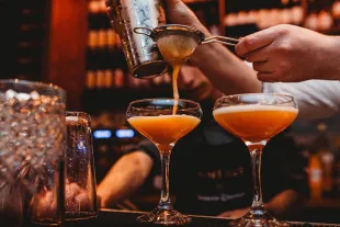 Gros plan sur un barman versant un cocktail orangé à travers une passoire dans deux coupes élégantes. En arrière-plan, l'ambiance tamisée d'un bar martiniquais avec des étagères de bouteilles de rhum.