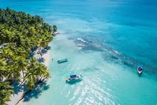 Vue aérienne d'une plage paradisiaque bordée de cocotiers et de bateaux dans une eau turquoise.